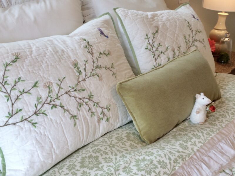 primary bedroom designed for comfort and relaxation, spacious bedroom oasis, bed layered with extra pillows, green accents create a soothing feel, carved wood French headboard, embroidered pillow shams, embroidered cotton coverlet, velvet cushion, Maxine Whitehead design